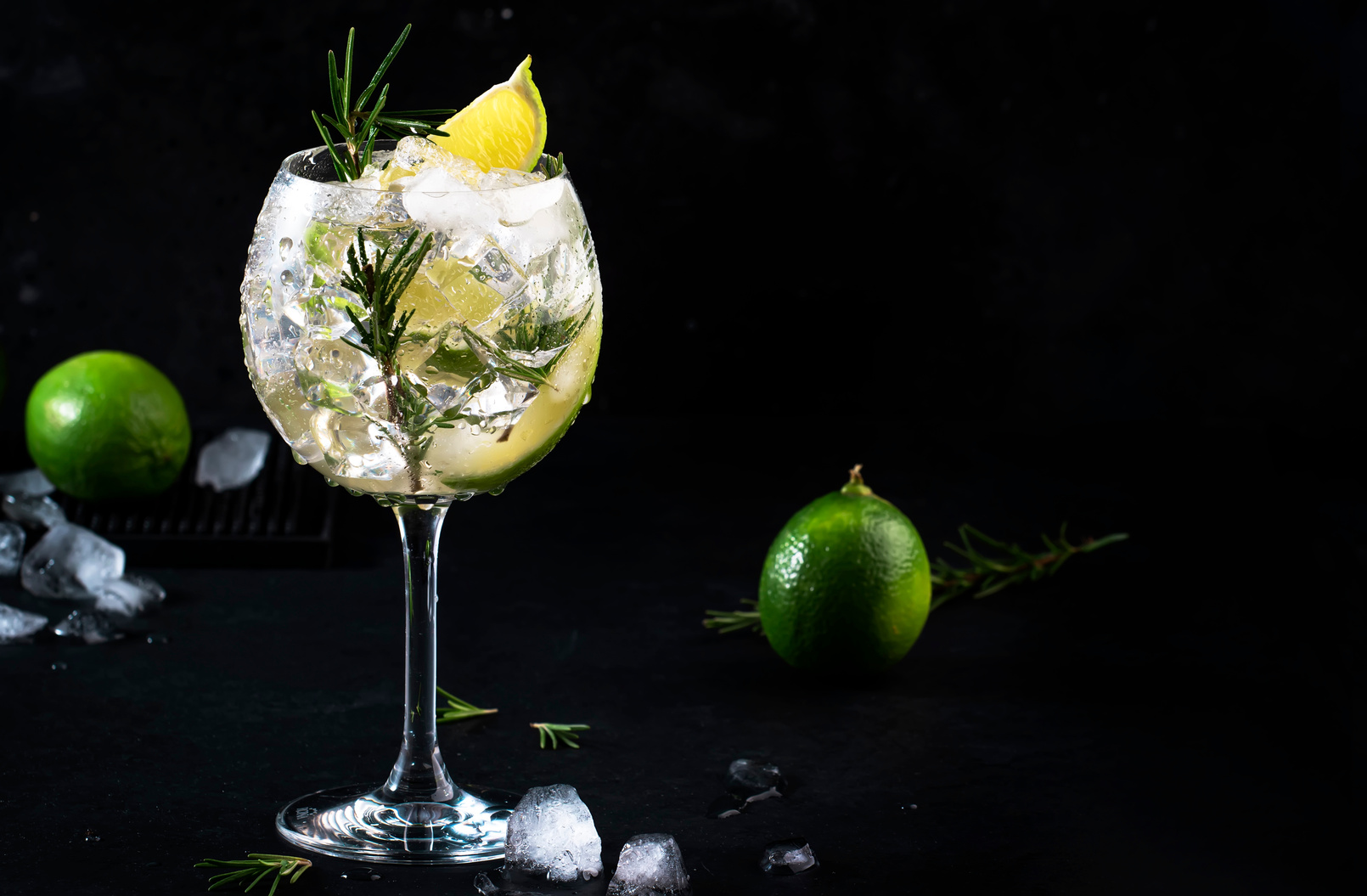 Gin tonic lime alcoholic cocktail drink with dry gin, rosemary, tonic and ice in big wine glass. Black bar counter background, steel bar tools